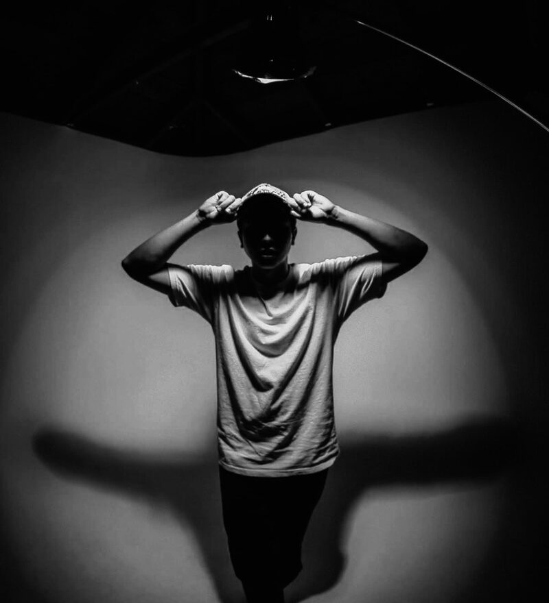 Vincent Broderick in a white T-shirt, hands on his cap, standing in a spotlight in black and white