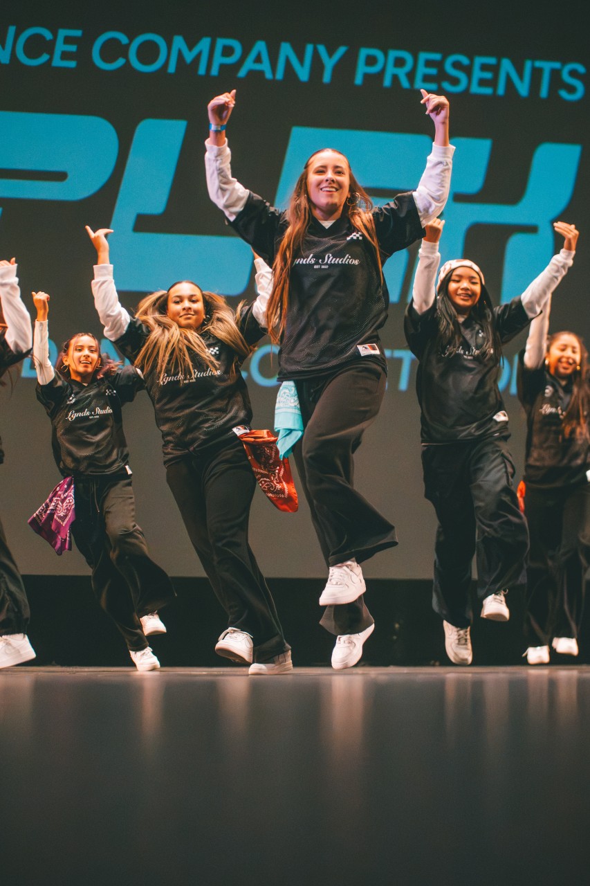 LGNDS dancers performing on stage with their arms raised in matching black jerseys.